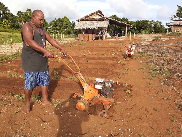 Man using a rotary hoe in a small field. A farm building is in the background